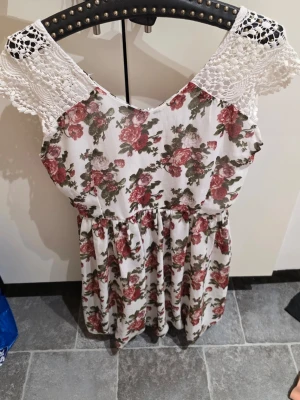 Blommig kortklänning från Club L - Supersöt kortklänning från Club L med vit botten och blommigt mönster i rött och grönt. Klänningen har spetsdetaljer vid axlarna och en lätt v-ringad hals. Materialet känns som bomull och polyester, perfekt för vår och sommar. Passformen är normal och klänningen är ärmlös.