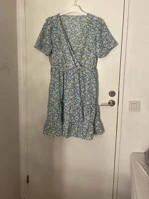 Blommig omlottklänning från Pigalle - Söt kort omlottklänning från Pigalle med blommigt mönster i blått, vitt och gult. Klänningen har volangkant nedtill, v-ringning och korta ärmar. Perfekt för sommarens alla tillfällen. Materialet är mjukt och lätt bomull. Pris kan diskuteras, tvätt ingår innan leverans ❤️ frågor kan ställas 