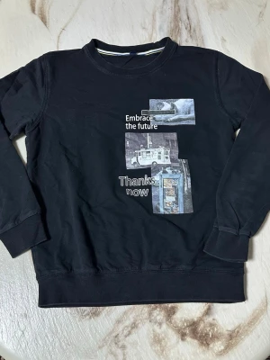 Svart sweatshirt med tryck, storlek S - Svart sweatshirt med coolt grafiskt tryck framtill som visar bilder och texten 'Embrace the future' och 'Thanks now'. Tröjan har rund hals, ribbade muddar och är långärmad. Materialet känns som mjuk bomull, perfekt för chill dagar.