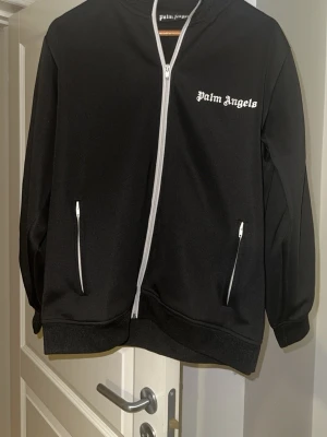 Svart trackjacka från Palm Angels - Palm Angels svart trackjacka med vita detaljer och dragkedjor. Jackan har vita ränder längs axlarna och bröstet pryds av Palm Angels-loggan. Tillverkad i polyester med ribbade muddar och två fickor med dragkedja framtill. Perfekt för en sportig och trendig look.