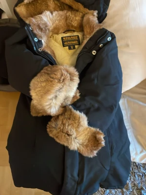 Svart barn dunjacka Woolrich med fuskpäls - Svart barn dunjacka från Woolrich med stor huva och mjuk fuskpäls i beige/brun ton på insidan och vid ärmsluten. Jackan har dragkedja och tryckknappar framtill. Perfekt för kalla vinterdagar och riktigt snygg stil.