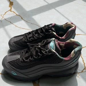 Svarta Nike Air Max Corteiz sneakers Aldrig Använda - Nike Air Max sneakers i svart och grått med turkosa detaljer. Ovandelen har mesh och skinn, och insidan har ett camouflage-mönster i grönt, beige och vinrött. Synlig Air Max-dämpning i sulan och snörning framtill. Snygg och unik design för dig som gillar streetstyle.