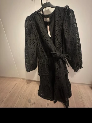 Svart Kelia midklänning med spets - Säljer en svart långklänning från Kelia (Nelly) i storlek 36. Klänningen har v-ringad omlottfront, långa ärmar och är dekorerad med broderad spets och hålmönster. Midjan markeras med ett knytband och kjolen har volangdetaljer. Passar perfekt till nyår eller annat firande, prislappen finns kvar. Nypris 899