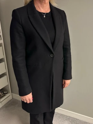 Svart kappa från Zara Woman - Svart kappa från Zara Woman i klassisk rak modell med en knapp framtill och diskreta fickor. Snygg krage och  ärmar med knappdetaljer vid ärmslut. Snygg både som öppen och stängd. Endast använt den ett par få gånger, så skicket är väldigt bra. Hör av er vid frågor eller mer bilder! 
