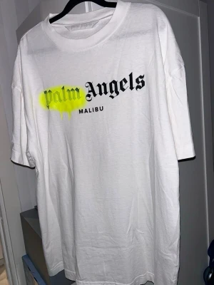 Palm Angels Malibu vit t-shirt XL - Palm Angels Malibu t-shirt i vitt med stor svart logotyp och neon-gul spraydetalj över 'Palm'. Klassisk rund hals och korta ärmar. Tillverkad i 100% bomull för skön och mjuk känsla. Perfekt för dig som gillar streetwear och vill sticka ut med en unik design.