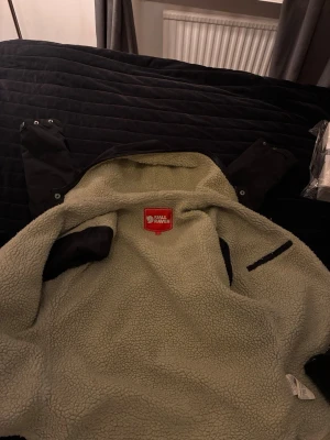 Fjällräven Greenland Winter Jacket – G-1000 – Teddyfodrad – Strl XS - Svart teddyjacka från Fjällräven i storlek XS med mjukt, beige teddyfoder och slitstark G-1000-ytterdel. Jackan har huva, tryckknappar och dragkedja samt klassisk Fjällräven-logga på ärmen. Perfekt för kalla dagar och outdoor vibes.
