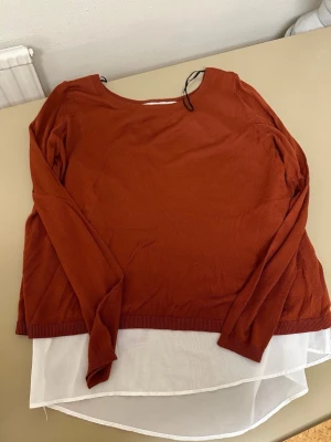 Rostbrun långärmad tröja med vit chiffong - Snygg rostbrun långärmad tröja med rund hals och vit chiffongdetalj som syns både fram och bak. Baksidan har en öppen design med omlott-effekt som visar det vita tyget. Perfekt för dig som gillar lager på lager och vill ha en unik look.