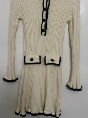 Vit ribbad stickad klänning med svarta detaljer - Säljer en vit ribbad stickad kläu med svarta kanter och knappar. Klänningen långärmad med volangkant, två dekorativa fickor framtill och svarta kontrastdetaljer längs knappraden och nederkant. Perfekt för dig som gillar klassisk och chic stil.