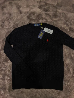 Svart kabelstickad tröja Polo Ralph Lauren - Svart kabelstickad tröja från Polo Ralph Lauren med klassisk rund hals och röd broderad logga på bröstet. Tröjan har lång ärm och ribbade muddar vid hals, ärmslut och nederkant. Perfekt för dig som gillar stilrena och tidlösa plagg.