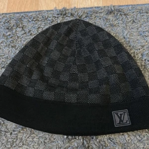 Svart rutig Louis Vuitton mössa - Snygg svart mössa från Louis Vuitton med diskret rutigt mönster och LV-logga framtill. Mössan är stickad och har en bred kant nedtill. Perfekt för dig som vill ha en lyxig accessoar med stilren design.