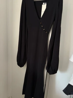 Svart långklänning från Adoore - Snygg svart långklänning från Adoore med v-ringning och dekorativa knappar framtill. Klänningen har långa puffärmar och en figurnära passform som ger en elegant siluett. Perfekt för dig som gillar stilrena och trendiga plagg.