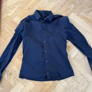 Mörkblå skjorta med knappar - Snygg mörkblå skjorta med klassisk krage och knappar hela vägen framtill. Skjortan har lång ärm och en stilren, enkel design som passar till många olika outfits. Materialet känns lätt och bekvämt mot huden. Perfekt för dig som gillar en clean look.