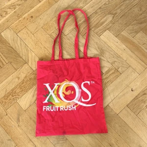 XQS Fruit Rush tote bag - Säljer en röd shoppingväska från XQS med trycket 'Fruit Rush' i vitt och färgglada detaljer. Väskan har långa handtag och är tillverkad i bomull, perfekt för att bära med sig till skolan eller stan. Enkel och snygg design med stor logga på framsidan.