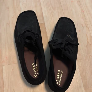 Svarta Clarks Originals Wallabee - Svarta Clarks Originals Wallabee i mocka med klassisk fyrkantig tå och snörning. Skorna har en platt crepesula och bruna innersulor med logga. Perfekta för dig som gillar tidlös och clean stil.