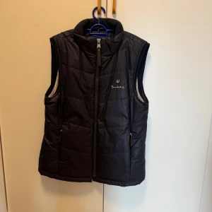 Svart quiltad dunväst från Bondelid - Marinblå dunväst från Bondelid i storlek L med quiltad design och hög krage. Västen har dragkedja framtill och två fickor med dragkedja. Perfekt för lager-på-lager och chill höst- eller vinterdagar. Klassisk look med diskret Bondelid-logga på bröstet.