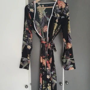 Blommig långklänning från Boohoo - Säljer en svart långklänning från Boohoo med färgglatt blommigt mönster i gult, orange, lila och grönt. Klänningen har lång ärm, vit kant längs öppningen och ett knytband i midjan. Perfekt för dig som gillar statement-plagg och vill sticka ut.