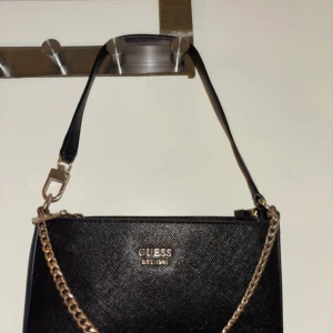 Svart axelväska från Guess med kedja - Snygg svart axelväska från Guess med guldfärgad kedjedetalj och logotyp framtill. Väskan har en stilren, rektangulär form och är tillverkad i skinnliknande syntetmaterial. Utrustad med dragkedja både på utsidan och insidan för smidig förvaring.