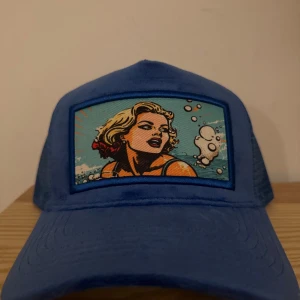 Blå keps med pop art-tryck från revenite - limiterad - Säljer en blå keps med coolt pop art-tryck framtill som visar en strandscen med en blond person och bubblor. Kepsen har böjd skärm och mesh på sidorna för extra ventilation. Perfekt för dig som vill sticka ut med en unik accessoar.