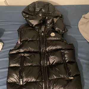 Svart dunväst från Moncler med huva - Svart, glansig dunväst från Moncler med stor, vadderad huva och två dragkedjeförsedda fickor. Västen har quiltad design och klassisk Moncler-logga på bröstet. Perfekt för lager-på-lager och streetwear. Dragkedja framtill och coola detaljer invändigt.