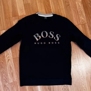 Svart sweatshirt från Hugo Boss - Svart sweatshirt från Hugo Boss med stort silverfärgat BOSS-tryck på bröstet. Klassisk rund halsringning med grå detalj på insidan av kragen. Långärmad och i mjukt material, perfekt för en stilren och avslappnad look.