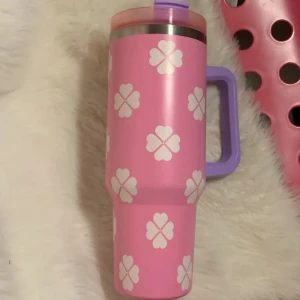 Pink Tumbler Mug with Straw and Handle - Söt rosa termosmugg med vita fyrklöver, lila handtag och lock samt sugrör. Perfekt för varma eller kalla drycker. Muggen ser ut att vara i gott skick utan synliga skador. Praktisk och trendig design för dagligt bruk.
