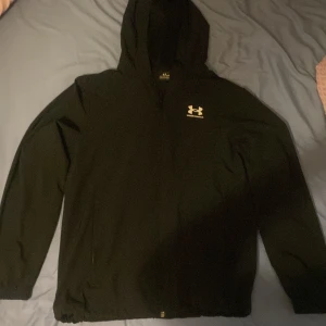 Svart vindjacka från Under Armour XS - Svart vindjacka från Under Armour i storlek XS. Jackan har huva, dragkedja framtill och elastiska muddar vid ärmslut och nederkant. Tillverkad i lätt polyester som andas, perfekt för aktiva dagar. Diskret logga på bröstet för en sportig vibe.