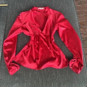 Julblus💕 - Säljer en röd blus från NA-KD i glansigt satinmaterial. Blusen har lång ärm, v-ringad hals och snygg twistad detalj framtill. Passformen är normal och tyget är mjukt och följsamt. Perfekt att styla med jeans eller kjol för en trendig look. Passar även xs/s se bilderna🙂