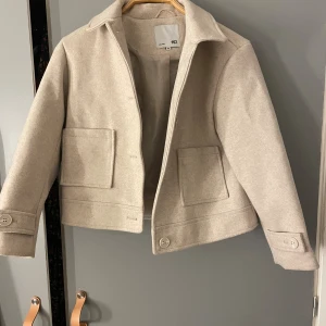 Beige ulljacka från 157 - Snygg beige jacka från 157 med klassisk krage och stora fickor framtill. Jackan är tillverkad i ull och har knappar vid ärmslut och nederkant. Perfekt för dig som gillar stilrena och enkla plagg med lite boxig passform.