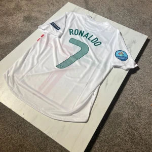 Portugal Ronaldo 7 Euro 2012 tröja Nike - Portugal fotbollströja från EM 2012 med Ronaldo 7 på ryggen. Vit med gröna och röda korsade ränder, broderat landslagsemblem och Nike-logga. Kortärmad modell i lätt och ventilerande Dri-FIT material. Patchar på ärmarna för Euro 2012 och Respect.
