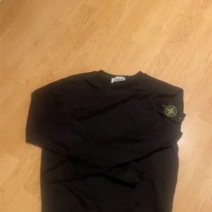 Svart långärmad tröja Stone Island - Svart långärmad tröja från Stone Island med klassisk rund hals och ikonisk logotyp-patch på vänster ärm. Tröjan är i mjuk bomull och har en clean, stilren look som passar perfekt till jeans eller cargo. Enkelt att matcha och riktigt snygg.