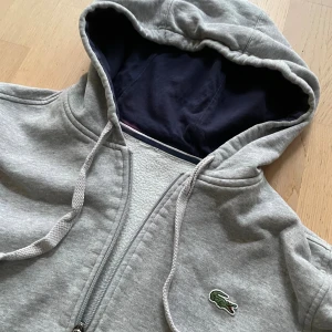 Grå hoodie från Lacoste med dragkedja - Snygg grå hoodie från Lacoste med klassisk krokodillogga på bröstet.  Har en liten fläck på ryggen därför de bra priset