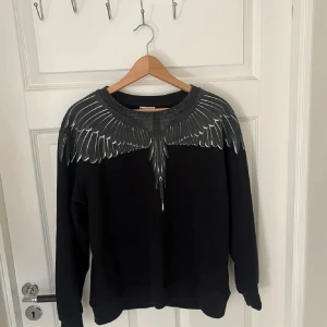 Svart sweatshirt Marcelo Burlon med vingar - Svart sweatshirt från Marcelo Burlon County of Milan med grått vingmönster över axlar och halsringning, både fram och bak. Tröjan har rund hals och långa ärmar, tillverkad i mjuk bomull. Perfekt statement-plagg för dig som gillar streetwear.