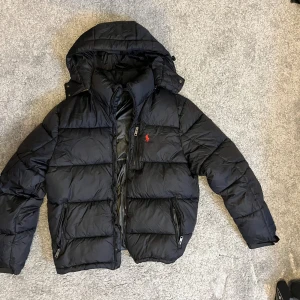 Svart dunjacka Polo Ralph Lauren M från Boozt - Köpt på Boozt! Sparsamnr använd köptens förra vintern. Säljer en svart pufferjacka från Polo Ralph Lauren i storlek M. Jackan har huva, två dragkedjefickor och klassisk röd Polo-logga broderad på bröstet. Fylld med dun för extra värme och har en clean, sportig look. Perfekt för kalla dagar.