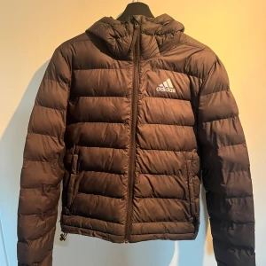 Svart pufferjacka från Adidas - Säljer en svart pufferjacka från Adidas med huva och dragkedja framtill. Jackan har Adidas-logga på bröstet och är vadderad för extra värme. Perfekt för kyliga dagar och har en clean, sportig look.