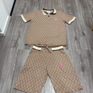 Bruna mönstrade pike & shorts från Casablanca - Säljer ett par bruna shorts & pike från Casablanca med beige och brunt rutmönster. De har snörning i midjan och en vit-beige rand upptill. Materialet känns mjukt och bekvämt, perfekt för chill dagar. På ena benet finns en röd logotyp-patch som ger extra stil. Storleken passar 164-176 piset kan diskuteras