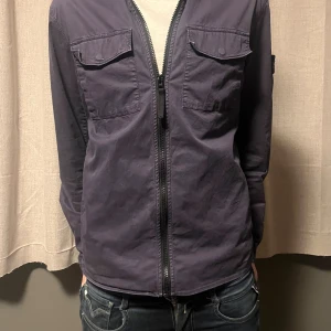 Mörkblå overshirt från Stone Island - Snygg mörkblå overshirt från Stone Island med klassisk patch på ärmen. Jackan har två bröstfickor med lock och knappar, samt dragkedja framtill. Materialet är mjukt och lätt, perfekt för lager-på-lager. Passar dig som gillar stilren och urban look. Storlek 170 men passar XS också.