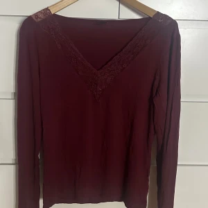 Vinröd långärmad topp med spets - Snygg vinröd långärmad topp med v-ringning och dekorativ spets längs halsringningen och axlarna. Tunn och mjuk, perfekt för dig som gillar stilrena detaljer och en mörk färgton. Passar dig som vill ha en enkel men ändå elegant look.