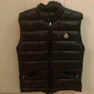 Svart dunväst från Moncler - Snygg svart dunväst från Moncler med quiltad design och hög krage. Väldigt fint skick! Storlek 5 passar perfekt på mig som är 175-180cm och väger 65-70kg 