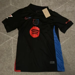 FC Barcelona Lamine Yamal Nike tröja - Säljer min helt nya/oanvända FC Barcelona fotbollströja från Nike med Lamine Yamal och nummer 19 på ryggen. Tröjan är svart med röda och blå detaljer, korta ärmar och tillverkad i Dri-FIT material för bästa komfort. Klubbmärke och sponsortryck på bröstet, UNHCR-logga på ryggen.