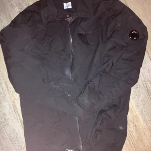 C.P. Company overshirt/skjorta - Svart overshirt från C.P. Company med klassisk krage, dragkedja framtill och en ikonisk linsdetalj på ärmen. Jackan har en bröstficka och är tillverkad i slitstark polyester. Perfekt för dig som gillar ett diskret plagg som drickat blickar och komplimanger. Funkar som tunnare höst overshirt eller till en sommaroutfit som skjorta! Storleken är M men sitter som S
