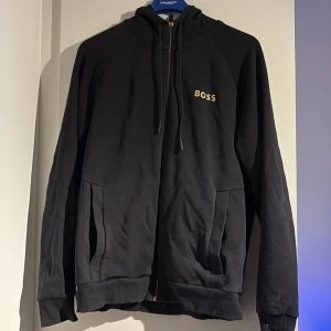 Svart hoodie från BOSS med dragkedja - Svart hoodie från BOSS i storlek M med guldfärgad BOSS-logga på bröstet. Tröjan har huva med snörning, två fickor fram och hel dragkedja. Ribbad mudd vid ärmslut och nederkant. Perfekt för chill eller streetwear.