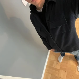 Mörkblå overshirt i ull, stl S - Säljer en stilren mörkblå overshirt i ull med klassisk krage och stora bröstfickor. Skjortan har knäppning framtill och långa ärmar, perfekt att slänga över en t-shirt eller hoodie för en chill look.