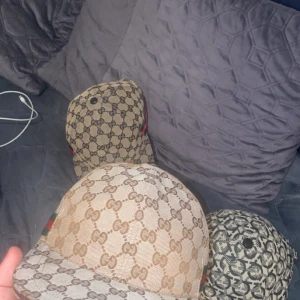 Gucci keps med GG-mönster och rödgrön detalj - Säljer en beige keps från Gucci med klassiskt GG-monogram över hela kepsen och rödgrön rand på sidan. Kepsen har böjd skärm och justerbar rem baktill i brunt läder. Perfekt för dig som vill sticka ut med en lyxig och ikonisk accessoar.