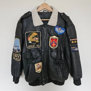Svart skinn bomberjacka med patchar - Riktigt fet svart vintage bomberjacka i skinn från Dual Control med US Army Airforces militärpatchar och en vit shearling krage. Jackan är illverkad under 80-talet och är mycket bra vintage skick, bara lädret är lite slitet. 