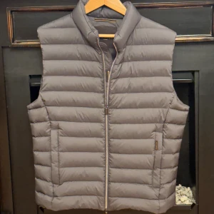 Grå dunväst från MooRER Verona - Snygg grå dunväst från MooRER Verona med quiltad design och hög krage. Västen har dragkedja framtill med märkta zip-pullers och två sidofickor. Insidan har gulddetaljer och exklusiva etiketter. Perfekt för lager-på-lager under kyliga dagar.