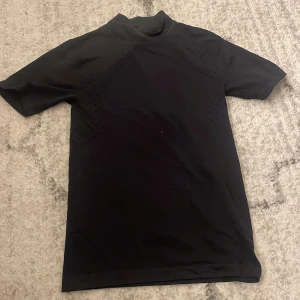 Svart t-shirt från JH Collection - Svart t-shirt från JH Collection med diskret mönster i tyget. T-shirten har korta ärmar och en lite högre krage för en clean look. Materialet känns mjukt och stretchigt, perfekt för en chill och stilren outfit.