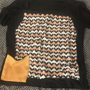 Svart blus med zigzag-mönster och ficka - Unik svart blus med korta ärmar och ett färgstarkt zigzag-mönster i orange, svart, grått och vitt på framsidan. Blusen har en cool orange ficka i hörnet som sticker ut och ger extra attityd. Materialet känns lätt och mjukt, perfekt för dig som gillar att sticka ut.