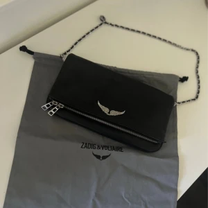 Svart axelväska från Zadig & Voltaire - Svart axelväska i skinn från Zadig & Voltaire med silverfärgad kedja och detaljer. Väskan har ett snyggt vingspänne framtill, flera dragkedjor och ett stilrent, rektangulärt format. Perfekt för dig som gillar minimalistisk design med en edgy touch.