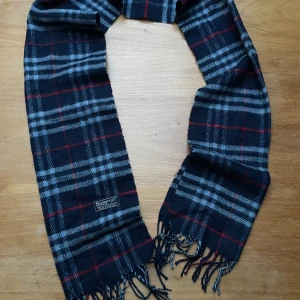 Rutig mörkblå halsduk från Burberrys - Snygg halsduk från Burberrys i klassiskt rutigt mönster med mörkblå, grå och röda toner. Gjord i 100% lammull, mjuk och varm med fransar i ändarna. Perfekt accessoar för att lyfta din vinterstil.