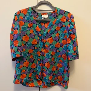 Färgglad blommig retro blus från Wahls strl C44 - Blommig retro blus från Wahls i storlek C44, med v-ringad hals och knappar framtill. Mönstret är färgstarkt med lila, turkos, orange och gröna toner. 